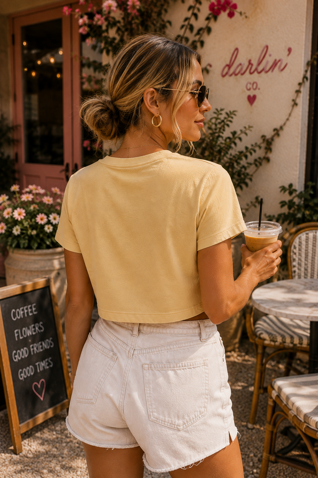A woman wearing a pale yellow cropped short-sleeve t-shirt and white denim shorts, photographed from behind in an outdoor cafe setting. She is holding an iced coffee and wearing sunglasses.