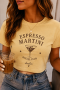 A woman wearing a cropped yellow t-shirt with a dark brown graphic that reads 'ESPRESSO MARTINI SOCIAL CLUB darlin''. She is holding an iced coffee and wearing blue jeans.
