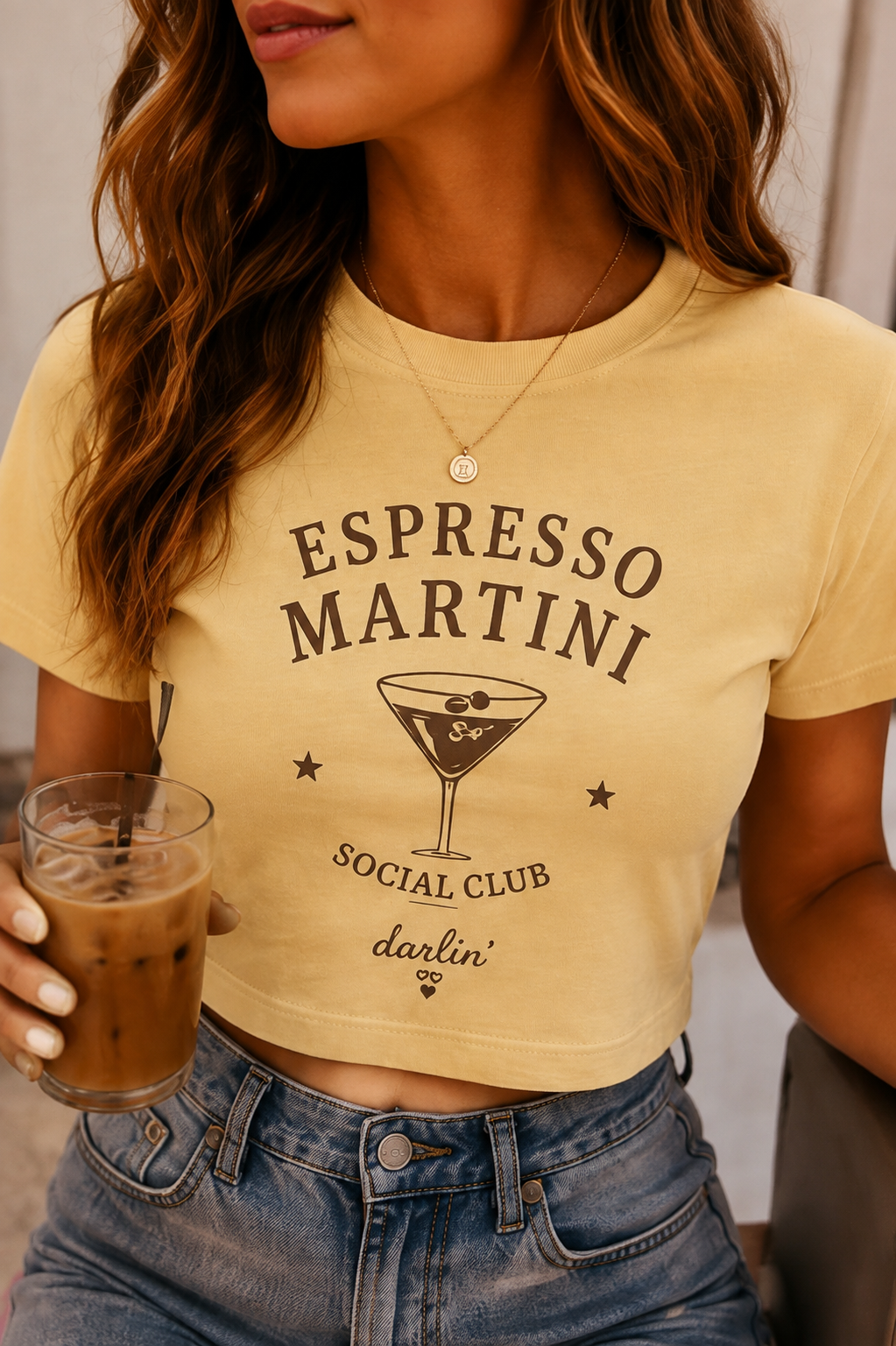 A woman wearing a cropped yellow t-shirt with a dark brown graphic that reads 'ESPRESSO MARTINI SOCIAL CLUB darlin''. She is holding an iced coffee and wearing blue jeans.
