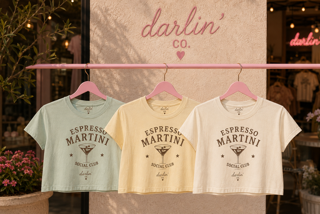 Three cropped short-sleeve T-shirts in pale mint, cream, and off-white hang on pink hangers from a pink clothing rack. The center cream shirt is most prominent, with printed text reading 'ESPRESSO MARTINI SOCIAL CLUB' and a martini glass graphic.