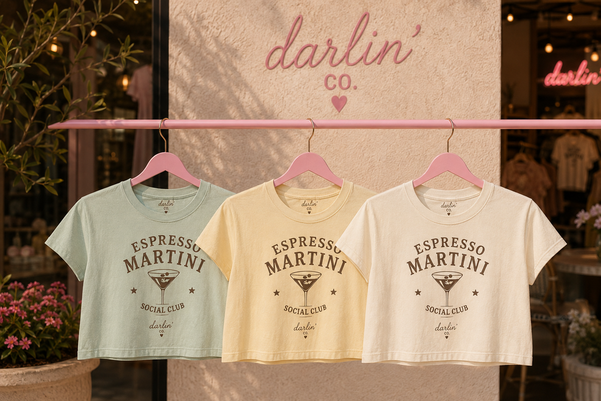Three cropped short-sleeve T-shirts in pale mint, cream, and off-white hang on pink hangers from a pink clothing rack. The center cream shirt is most prominent, with printed text reading 'ESPRESSO MARTINI SOCIAL CLUB' and a martini glass graphic.