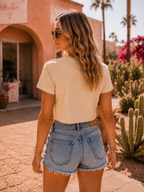 A woman wearing a beige short-sleeve crop top and light-wash denim shorts stands outdoors in a sunny desert town setting with cacti and a pink building in the background.