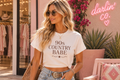 A woman wearing a white graphic t-shirt with '90s COUNTRY BABE' printed in brown text, styled with light-wash jeans and sunglasses, standing in front of a boutique wall with a pink neon sign.