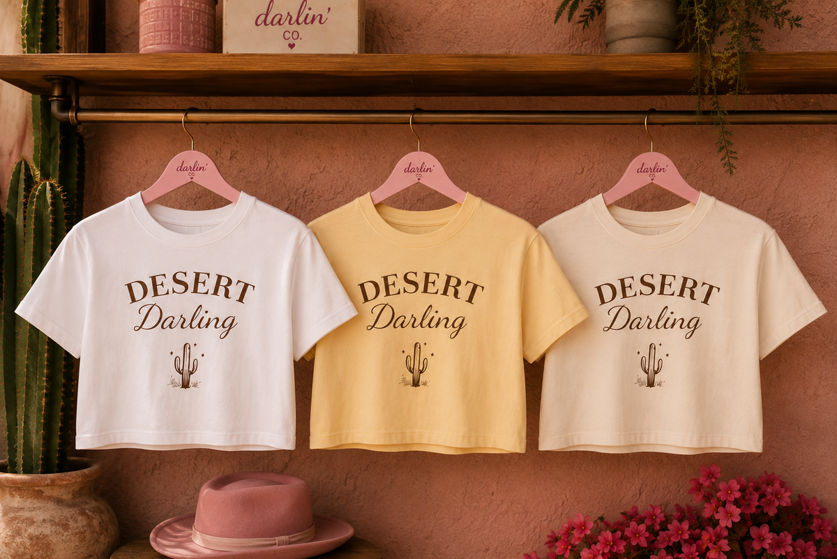 Three cropped graphic T-shirts hanging on pink hangers against a terracotta wall, with a wooden shelf and plants in the background. The shirts are white, pale yellow, and cream, each printed with the words 'DESERT Darling' and a small cactus graphic.