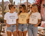 Three women standing outdoors in a desert setting wearing matching graphic crop T-shirts with the words 'DESERT Darling' and a small cactus illustration. The shirts come in white, pale yellow, and light blue-gray tones, paired with denim shorts.