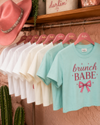 A row of short-sleeve graphic T-shirts hanging on pink hangers labeled Darlin Co, displayed on a clothing rack with a pink hat and cactus props in a warm retail setting. The front shirt is light aqua with 'brunch BABE' and a pink bow graphic.