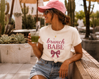 A woman sitting outdoors on a wooden bench at a cafe, wearing a pink baseball cap, sunglasses, a cream graphic t-shirt that reads 'brunch BABE' with a pink bow, and denim shorts while holding a green drink.