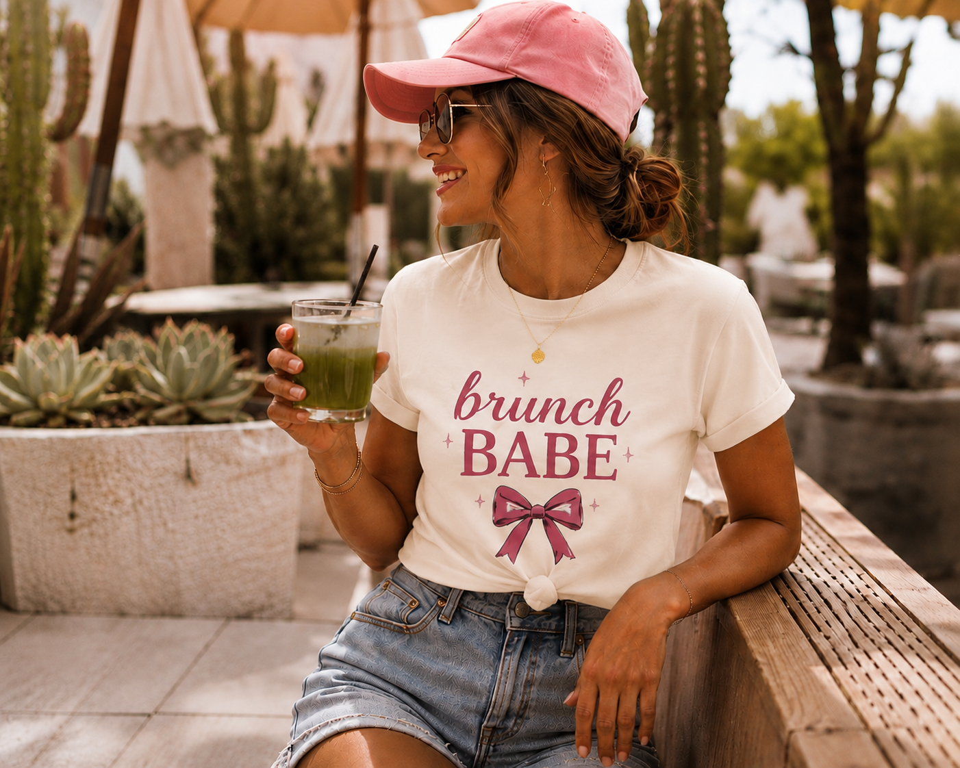 A woman sitting outdoors on a wooden bench at a cafe, wearing a pink baseball cap, sunglasses, a cream graphic t-shirt that reads 'brunch BABE' with a pink bow, and denim shorts while holding a green drink.