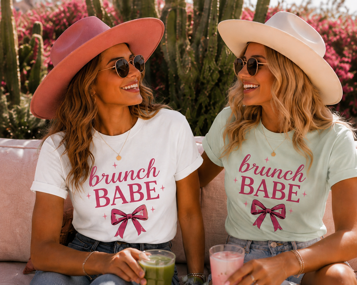 Two women sitting on a pink couch outdoors, wearing matching Brunch Babe graphic T-shirts with a bow illustration. The woman on the left wears a pink hat and sunglasses, and the woman on the right wears a cream hat and sunglasses.
