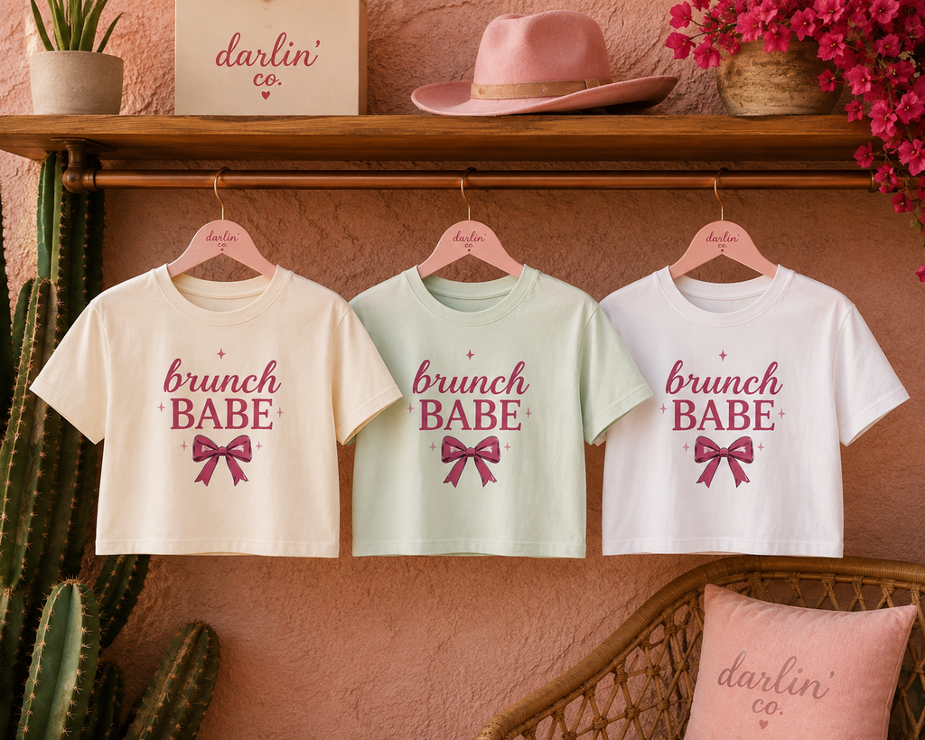 Three cropped short-sleeve T-shirts hanging on wooden hangers against a textured pink wall, with a wooden shelf and decor above and below. The shirts are cream, pale mint, and white, each printed with the phrase 'brunch BABE' in pink script and a pink bow graphic.