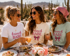 Three women sitting at a table outdoors in a desert setting, wearing matching Brunch Babe graphic T-shirts with a pink bow illustration. The shirts are white, cream, and light mint green, with saguaro cacti and mountains in the background.