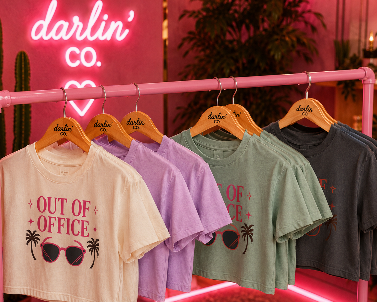 Four Darlin' Co. short-sleeve graphic t-shirts in cream, lavender, sage green, and charcoal gray hang on a pink clothing rack in front of a pink neon sign.