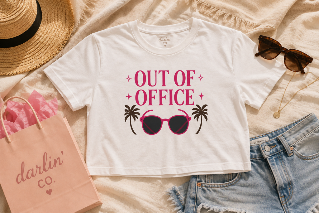 A white cropped graphic T-shirt laid flat on a beige fabric surface, surrounded by styled props including a straw hat, sunglasses, denim shorts, a pink shopping bag, and a necklace. The shirt graphic reads “OUT OF OFFICE” in pink with black palm trees and pink sunglasses.