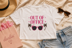 A white cropped graphic T-shirt laid flat on a beige fabric surface, surrounded by styled props including a straw hat, sunglasses, denim shorts, a pink shopping bag, and a necklace. The shirt graphic reads “OUT OF OFFICE” in pink with black palm trees and pink sunglasses.