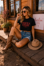 A woman sits on wooden steps in front of a rustic cabin, wearing a black graphic T-shirt that reads 'TEQUILA COWGIRL' in red letters, denim shorts, and brown cowboy boots. A tan cowboy hat rests beside her, and a sign reading 'Darlin' Co.