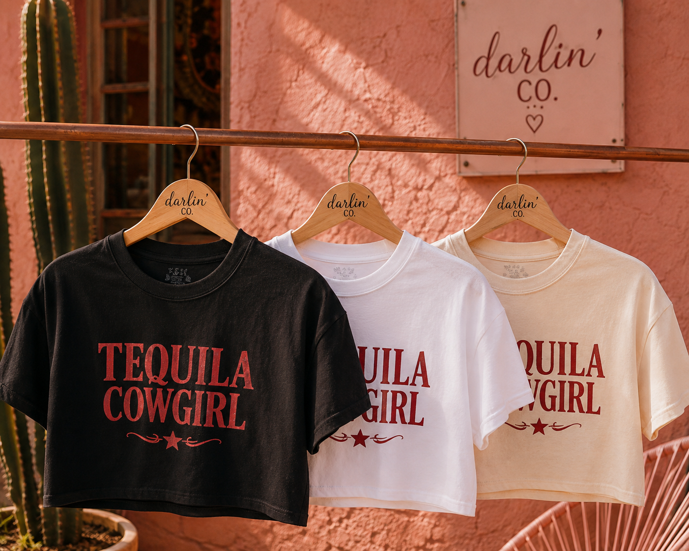 Three cropped graphic T-shirts hanging on wooden hangers against a textured pink wall. The shirts are black, white, and cream, each featuring the words 'TEQUILA COWGIRL' in a dark red serif font with decorative flourishes.