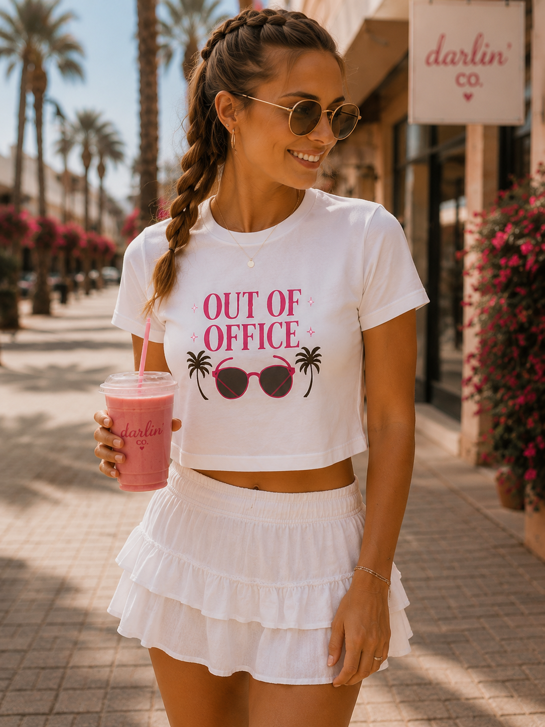 A woman wearing a white crop top t-shirt with a pink and black graphic that reads 'OUT OF OFFICE' with sunglasses and palm trees, paired with a white ruffled mini skirt. She is holding a pink drink on a sunny outdoor sidewalk.