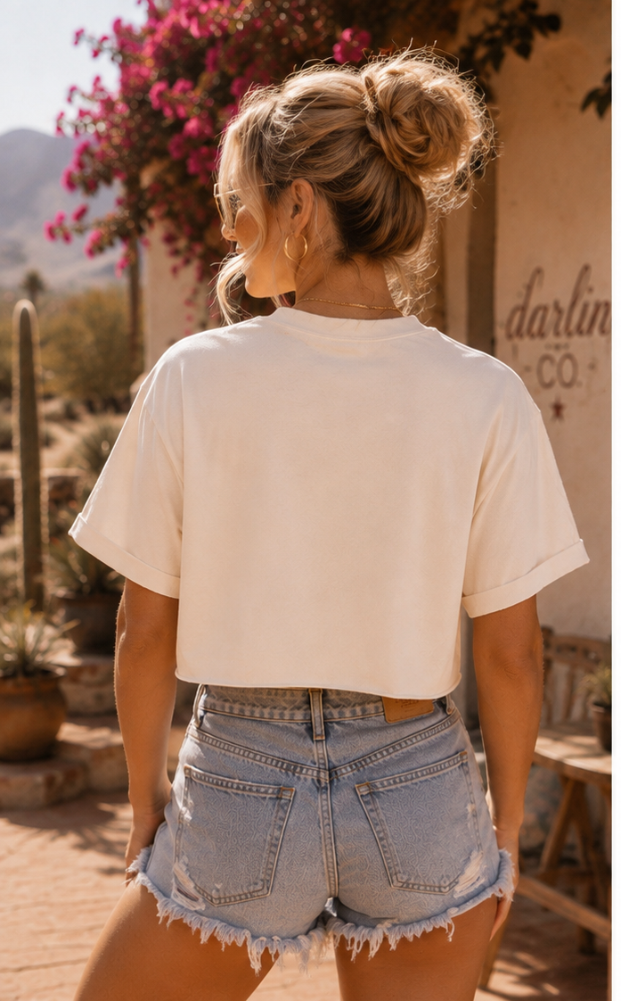 A woman viewed from behind wearing a cream-colored short-sleeve crop top and light-wash denim shorts in an outdoor desert setting.