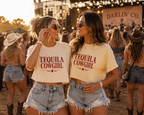 Two women at a sunset outdoor festival wearing matching cream-colored crop top t-shirts with 'TEQUILA COWGIRL' printed in red text, paired with denim shorts.