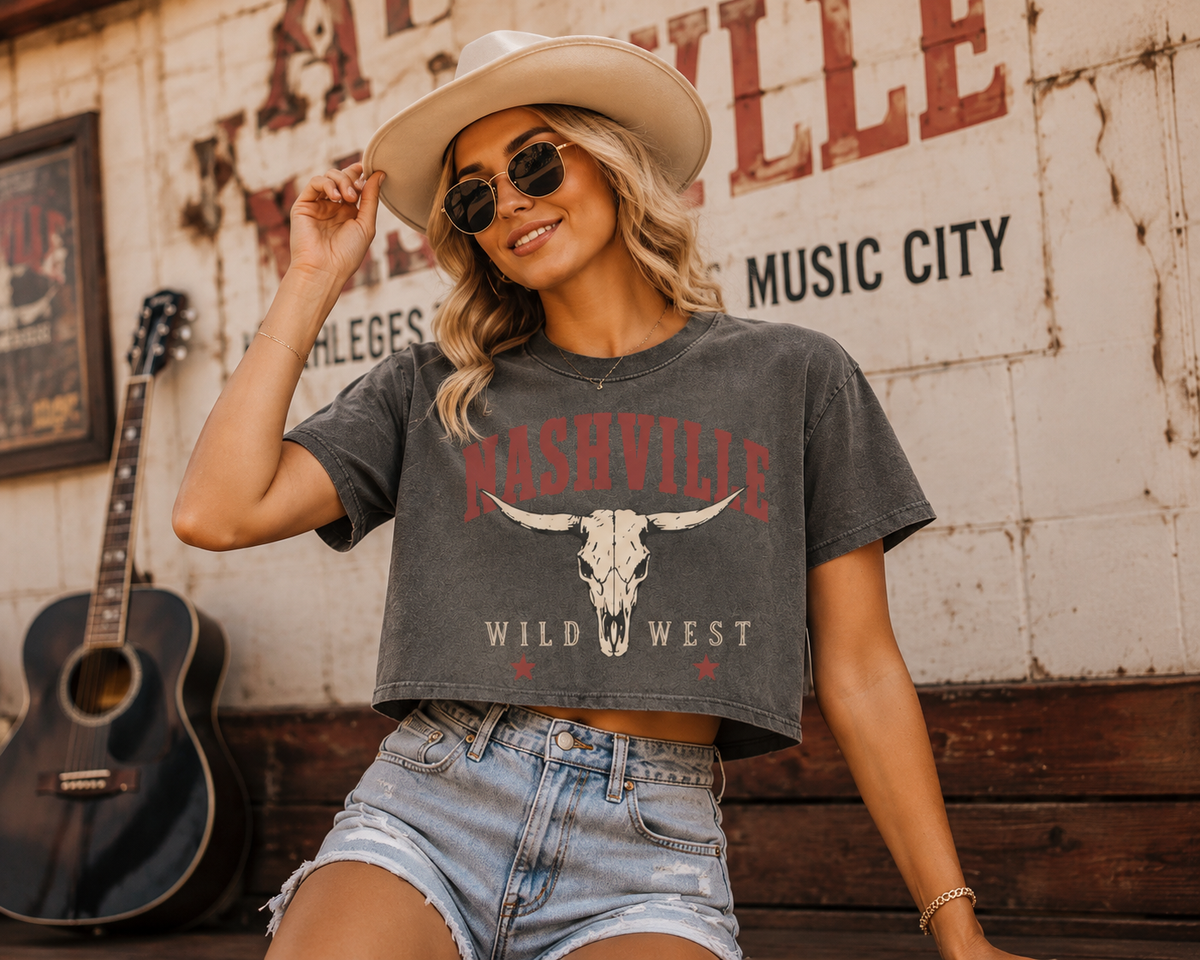 A woman wearing a Nashville Wild West graphic crop top, denim shorts, and a cowboy hat poses in front of a rustic wall with a guitar.