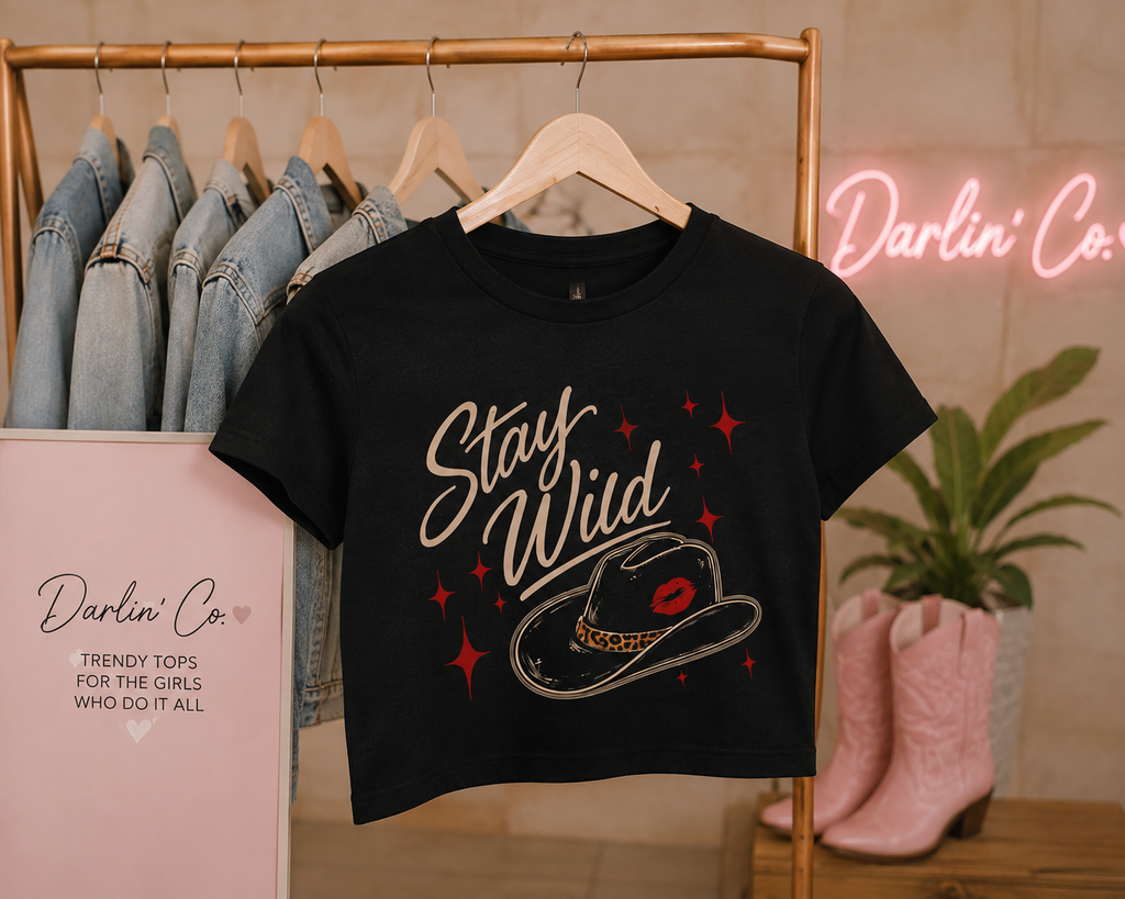 Black Stay Wild graphic crop tee on a wooden hanger, featuring beige script, cowboy hat with red lips, and red star accents in a Darlin’ Co. boutique setting.