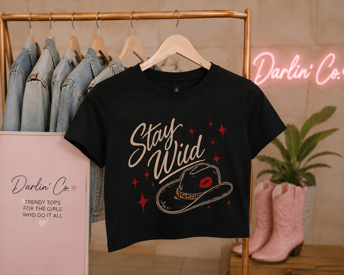 Black Stay Wild graphic crop tee on a wooden hanger, featuring beige script, cowboy hat with red lips, and red star accents in a Darlin’ Co. boutique setting.