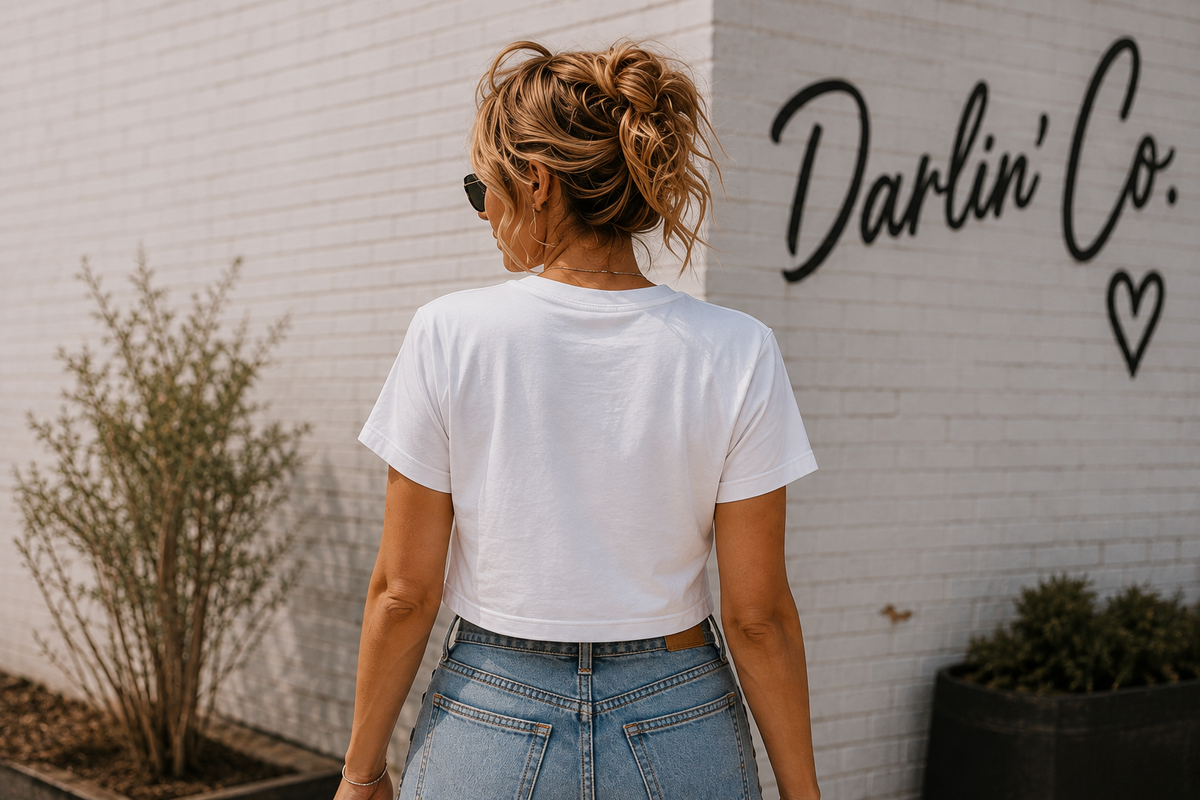 Back view of a woman wearing a white cropped tee with light blue jeans in front of a white brick wall featuring the Darlin’ Co. logo.