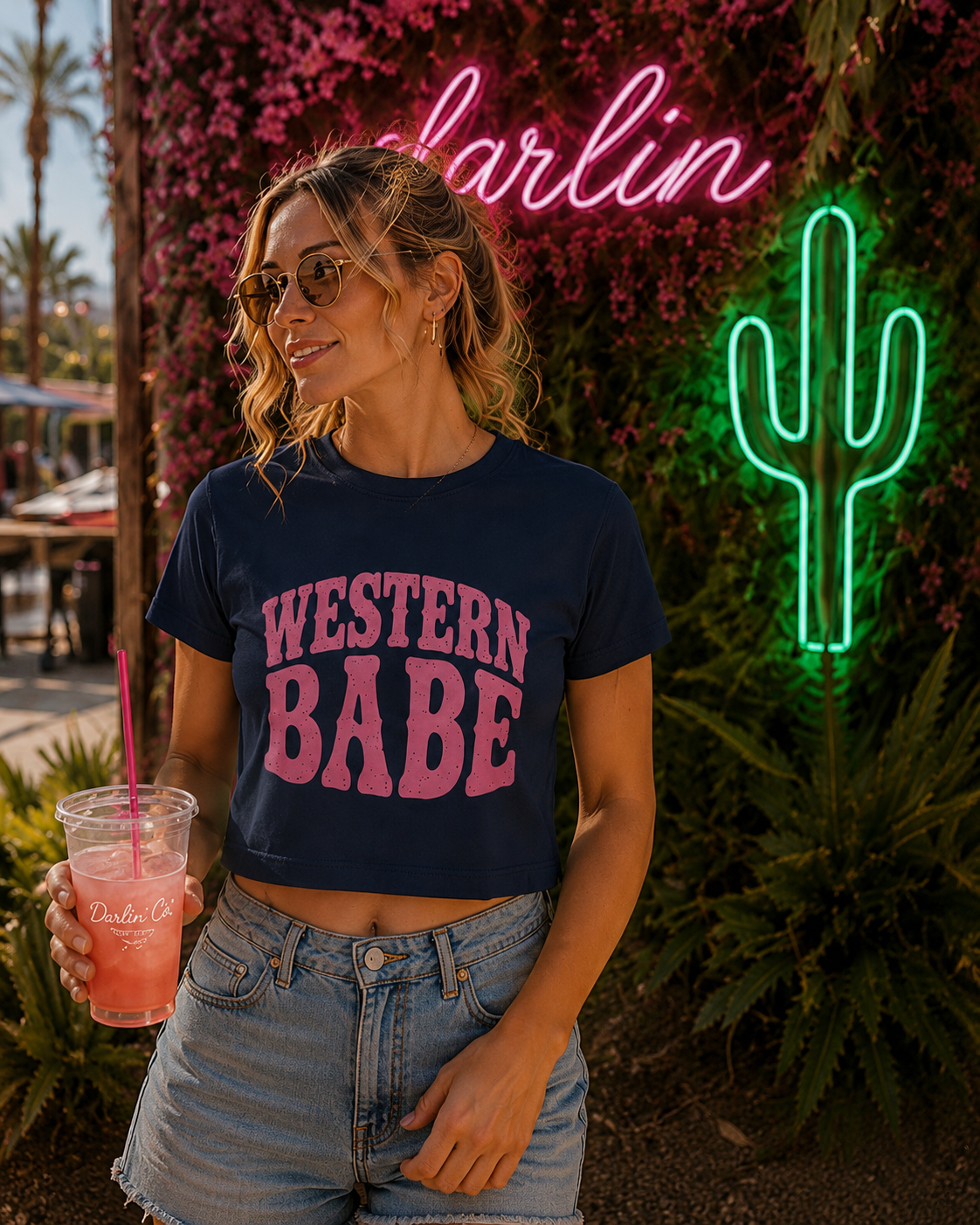 A woman wearing a navy blue cropped t-shirt with 'WESTERN BABE' printed in pink letters, paired with light-wash denim shorts. She is holding a pink drink and standing in front of a green neon cactus sign and a pink 'darlin' neon sign against a wall of pink flowers.