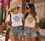 Two women standing outdoors in a desert-style storefront, wearing matching graphic crop T-shirts that read '90s COUNTRY BABE' in black text. One shirt is white and the other is a warm cream/beige.