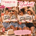 Four women in a rustic outdoor setting wearing matching white graphic tees that read 'HOWDY darlin' CO.' in black glitter-style lettering with a pink heart.